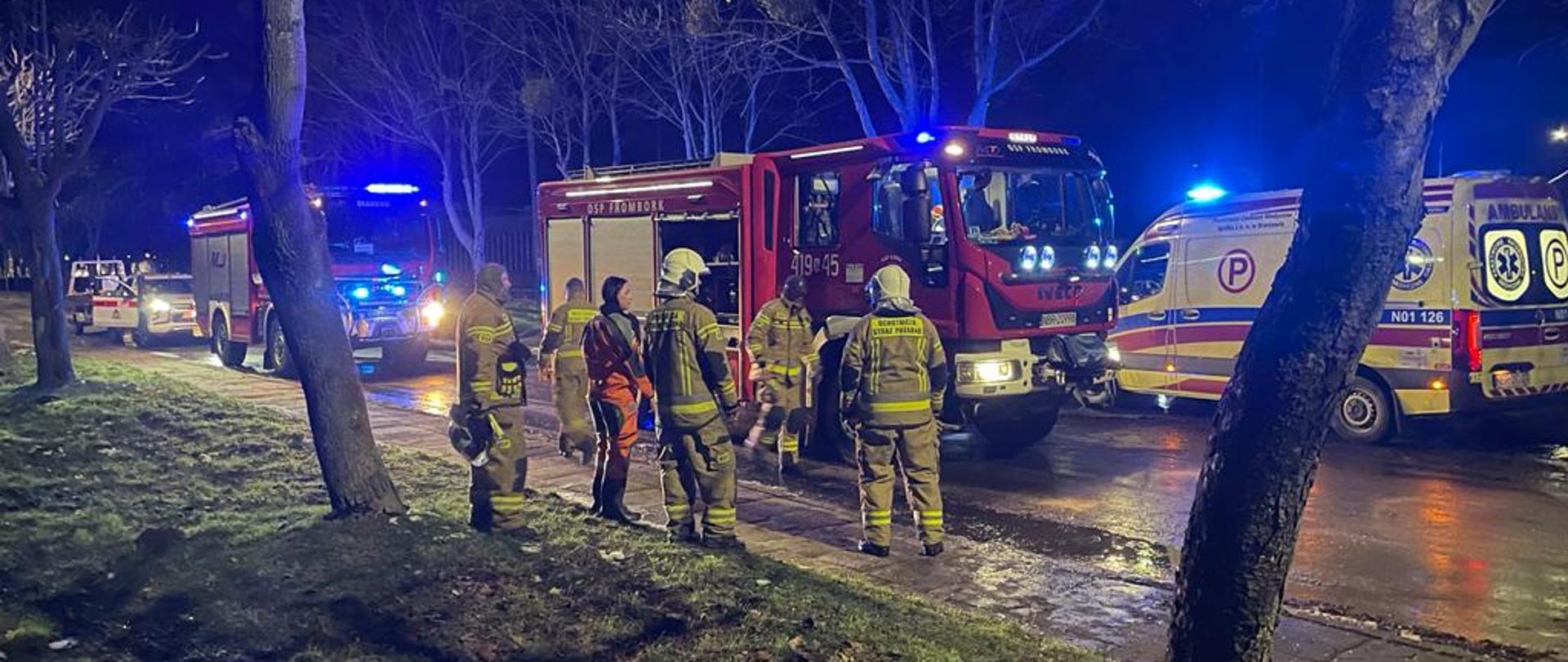 Zdjęcie zrobione nocą. Na dole zdjęcia zielony trawnik, dalej na drodze ustawiane samochody, dwa strażackie i karetka pogotowia. Przy samochodzie stoją strażacy. Samochody mają włączone niebieskie światła ostrzegawcze.