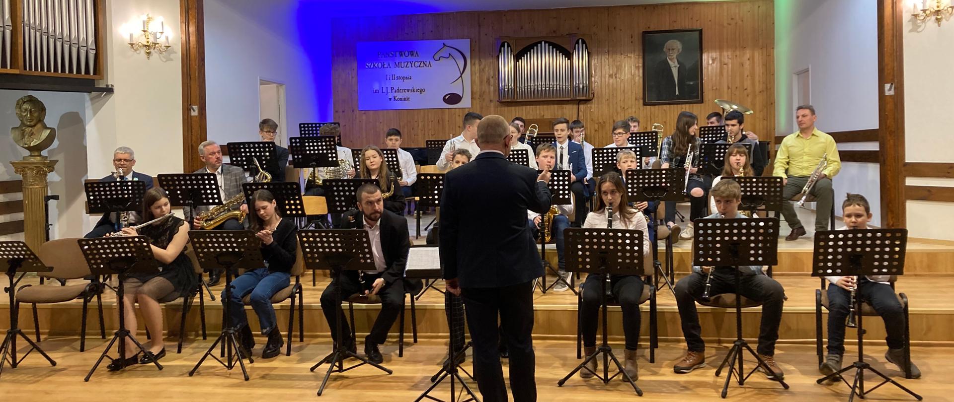Sala koncertowa PSM w Koninie, na scenie siedzi orkiestra szkolna, dyrygent stoi odwrócony plecami, muzycy trzymają instrumenty, w tle niebieskie światło