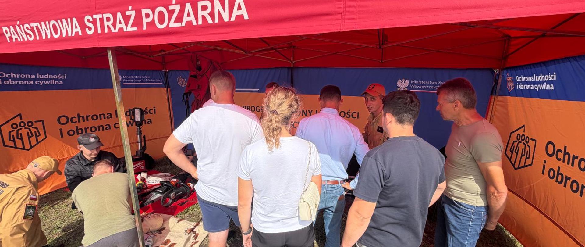 Podlascy strażacy #PSP i #OSP czynnie włączyli się w trening, przekazując informacje dotyczące bezpieczeństwa. Na zdjęciu strażacy przekazują treści dotyczące bezpieczeństwa, w tle namioty pod którymi leżą elementy wyposażenia plecaka ewakuacyjnego. Wszystkiemu przygląda się ludność cywilna. Całość odbywa się na podwórku. 