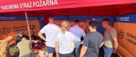 Podlascy strażacy #PSP i #OSP czynnie włączyli się w trening, przekazując informacje dotyczące bezpieczeństwa. Na zdjęciu strażacy przekazują treści dotyczące bezpieczeństwa, w tle namioty pod którymi leżą elementy wyposażenia plecaka ewakuacyjnego. Wszystkiemu przygląda się ludność cywilna. Całość odbywa się na podwórku. 