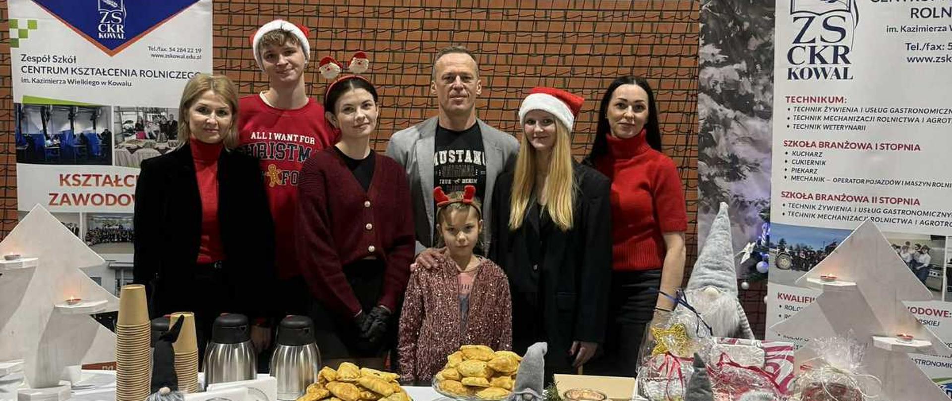 Zdjęcie przedstawia ośmiu uśmiechniętych ludzi, w tym siedmiu dorosłych i jedno dziecko, stojących za stołem z dekoracjami i potrawami świątecznymi. Osoby te wydają się uczestniczyć w wydarzeniu o charakterze bożonarodzeniowym, prawdopodobnie kiermaszu lub spotkaniu wigilijnym, zorganizowanym przez Zespół Szkół Centrum Kształcenia Rolniczego (ZSCKR) im. Kazimierza Wielkiego w Kowalu, na co wskazują transparenty widoczne w tle.