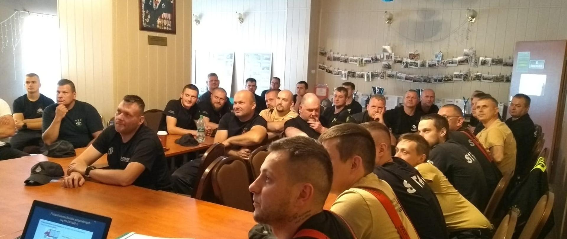 Zdjęcie wykonano w pomieszczeniu świetlicy budynku Ochotniczej straży pożarnej w Sępólnie Krajeńskim. Strażacy Ochotniczej Straży Pożarnej ubrani w mundury koszarowe siedzą przy stołach słuchając wykładu. 