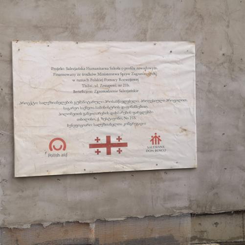 Tablica informacyjna na temat projektu, na której napisane jest: Projekt: Salezjańska Szkoła Humanitarna o profilu Zawodowym. Finansowany ze środków Ministerstwa Spraw Zagranicznych w ramach Polskiej Pomocy Rozwojowej. Napisy po polsku i po gruzińsku. Na dole logo Polskiej pomocy, flaga Gruzji i logo Salezian Don Bosco. 