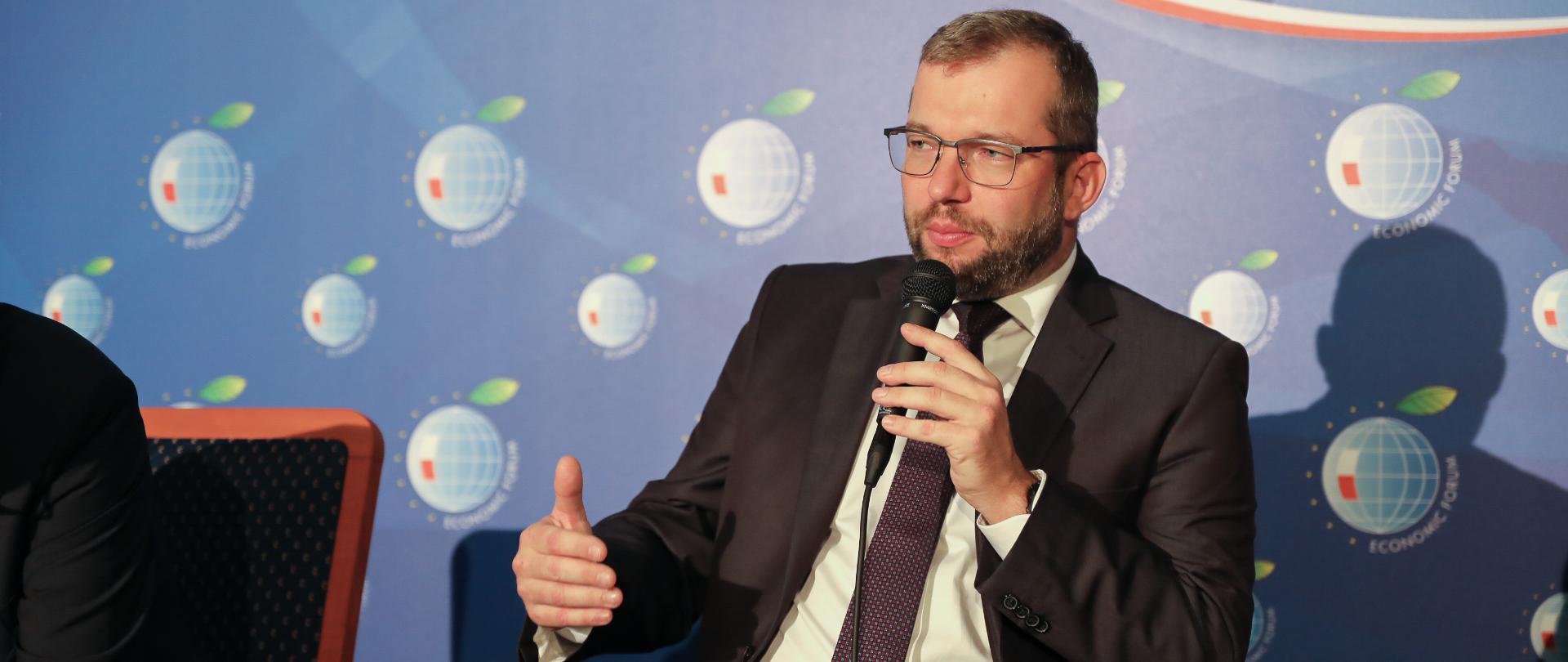 Minister Puda siedzi na fotelu, trzyma w lewym ręku mikrofon i mówi do niego.