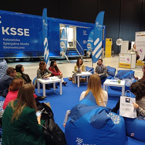 Grupa osób siedzi na niebieskich pufach w okręgu. Z tyłu widać samochód z napisem Katowicka Specjalna Strefa Ekonomiczna