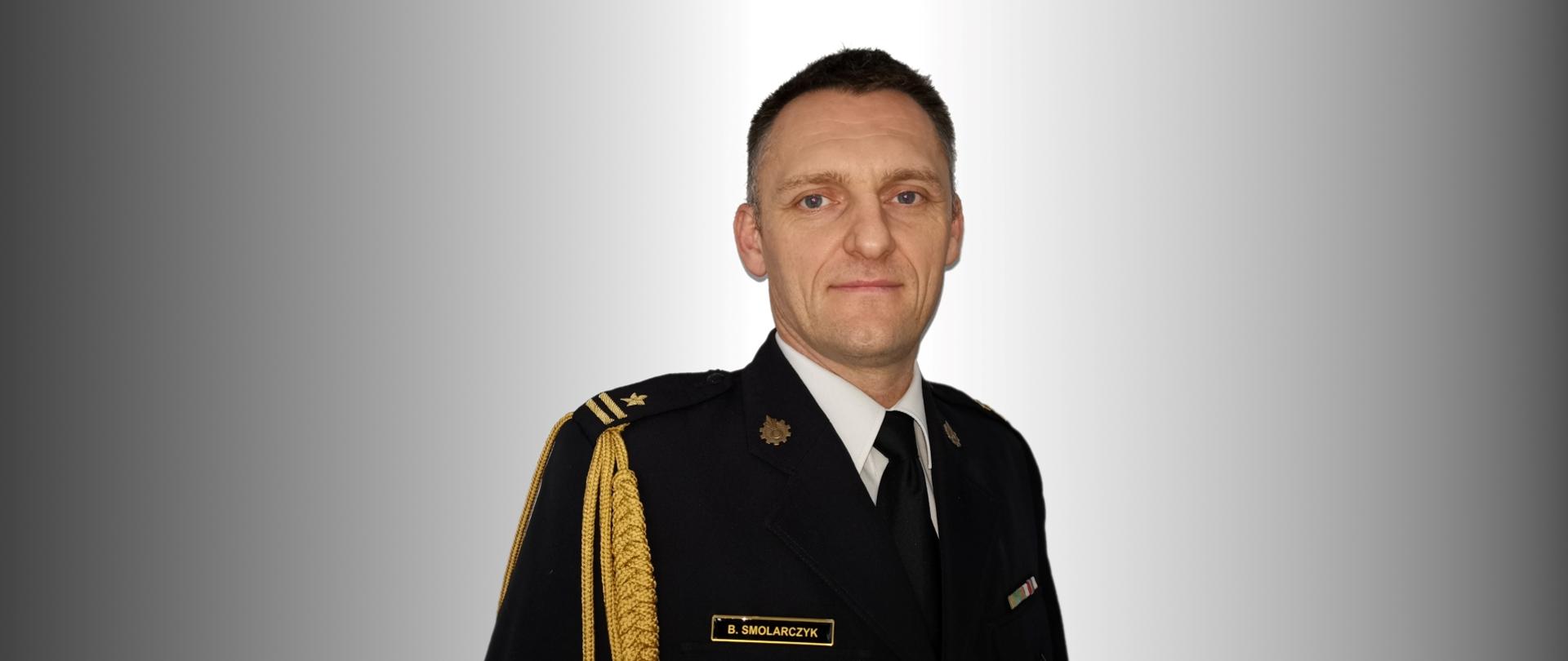 Zdjęcie przedstawia mł. bryg. Bartosza Smolarczyka w mundurze galowym na biało-szarym tle.
