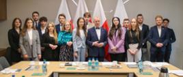 Zdjęcie zbiorowe, grupa młodzieży stoi w pomieszczeniu ze stołami ustawionymi w czworokąt, za nimi cztery polskie flagi.