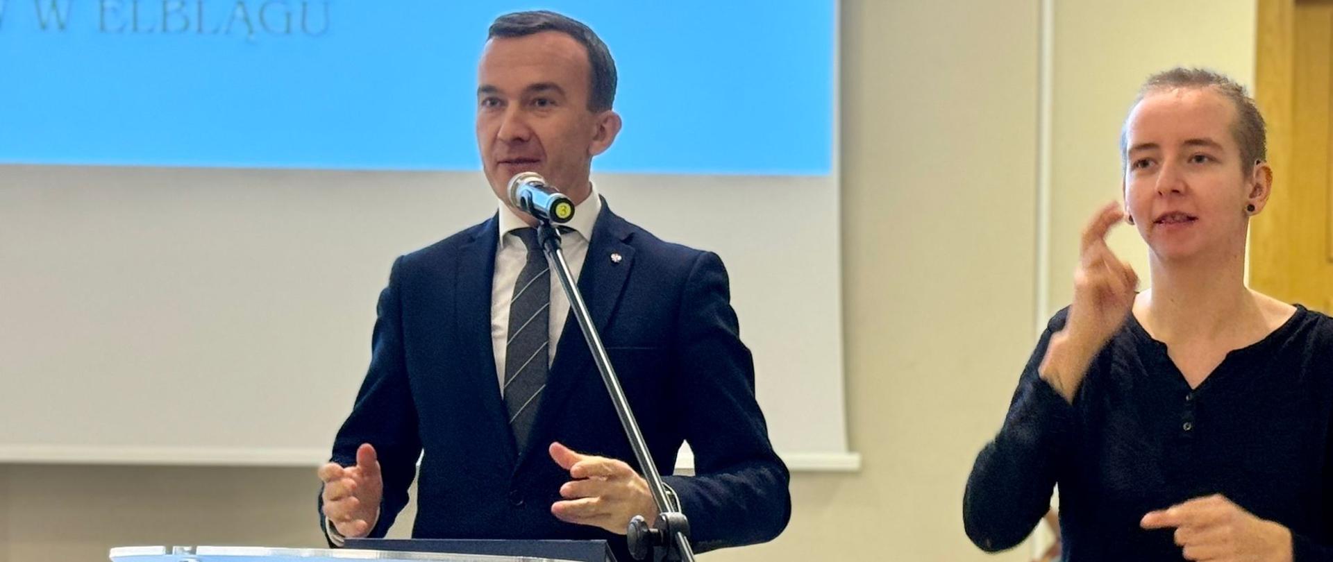 Mężczyzna w granatowym garniturze przemawia z mównicy 