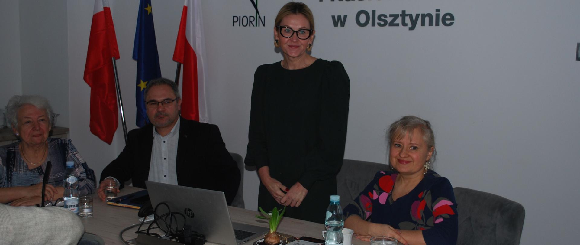 przy stole od lewej siedzi kobieta, mężczyzna, pośrodku stoi kobieta przemawiająca do uczestników a po jej lewej stronie siedzi jeszcze jedna kobieta