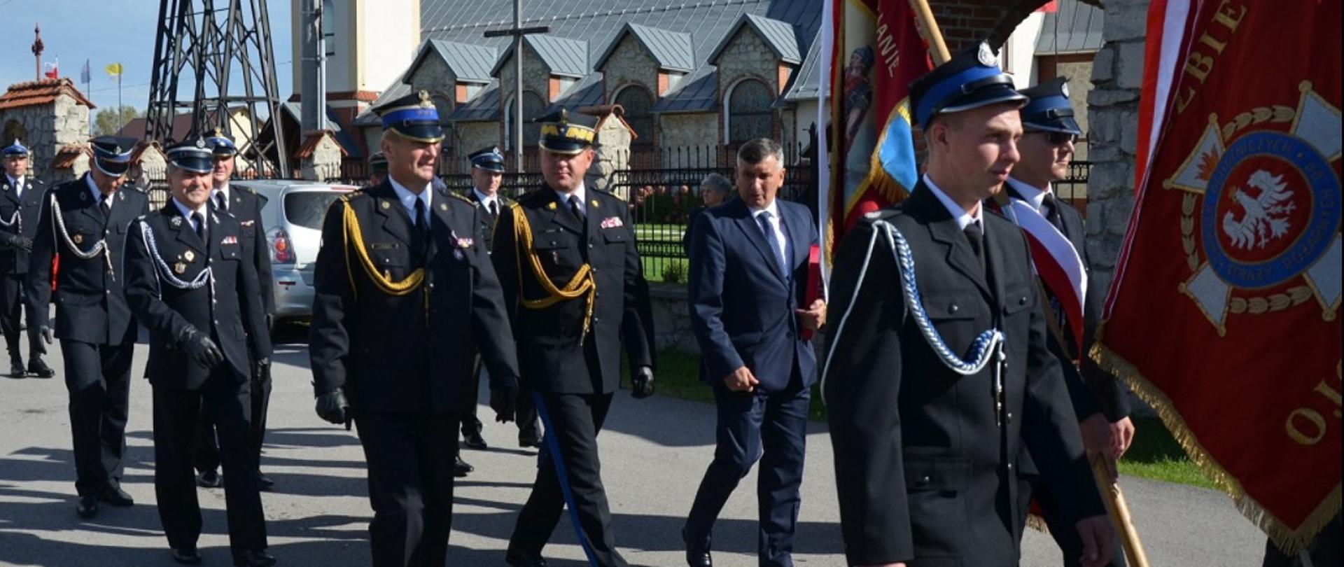 Na zdjęciu znajdują się maszerujący strażacy w mundurach galowych na czele których podąża sztandar jednostki z asystą.