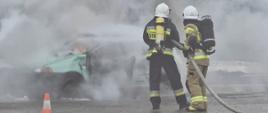 Dwóch strażaków w mundurach specjalnych gasi płonący samochód. Wokół samochodu dużo pary wodnej. 