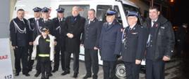 Zebranie sprawozdawcze OSP Ostoje. Plac przed remizą OSP. Na tle busa strażackiego koloru białego stoją przedstawiciele OSP wraz z ministrem Dziedziczakiem oraz zastępcą komendanta powiatowego PSP.