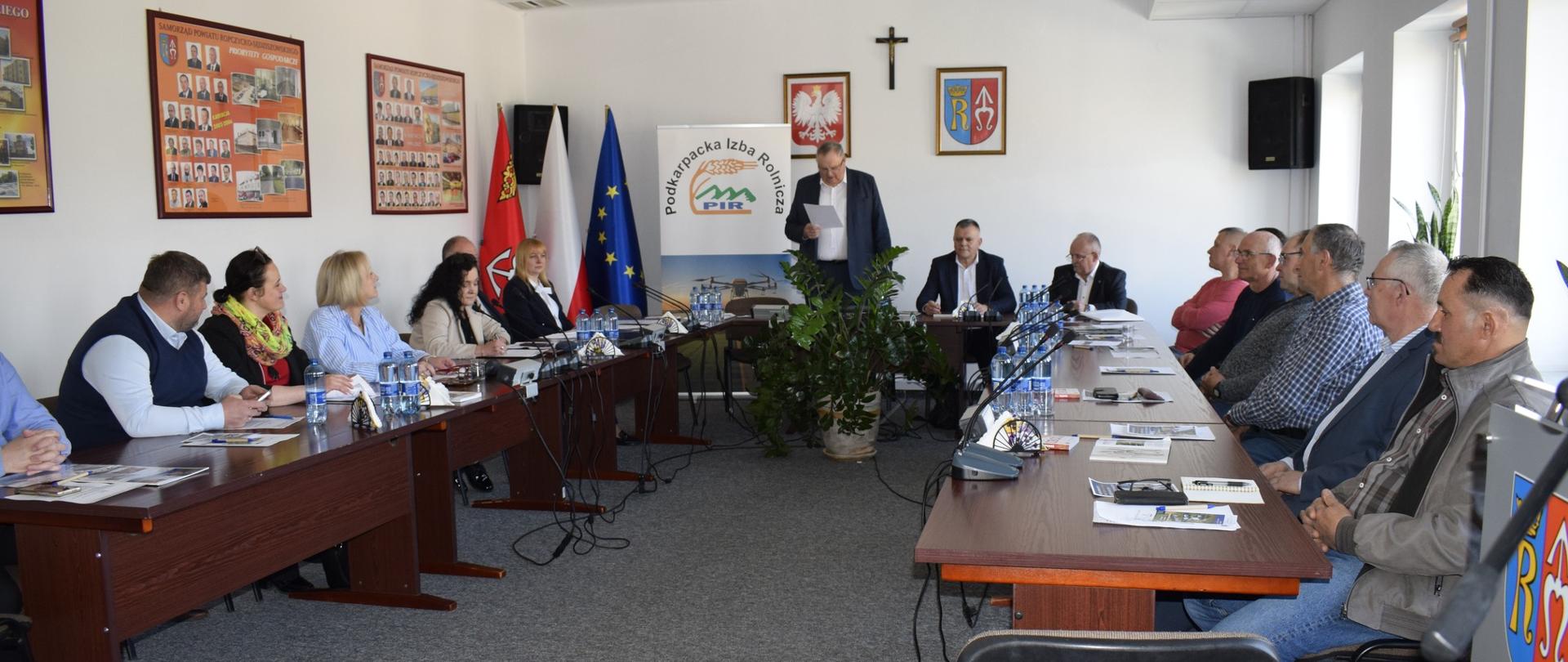 Szerokie ujęcie sali konferencyjnej podczas oficjalnego spotkania. W pomieszczeniu ustawione są drewniane stoły w kształcie podkowy, przy których siedzi kilkanaście osób – kobiety i mężczyźni w średnim wieku, ubrani w stylu półformalny. Na stołach znajdują się mikrofony, butelki z wodą, dokumenty i notesy. W centrum, na końcu stołów, stoi mężczyzna w ciemnej marynarce, który czyta z kartki i wygląda na osobę prowadzącą spotkanie. Za nim widoczny jest baner z napisem „Podkarpacka Izba Rolnicza (PIR)”, a obok stoją flagi Polski i Unii Europejskiej. Na ścianie wiszą godło Polski, herb oraz krzyż. W pomieszczeniu są jasne ściany, sufitowe lampy i okna po prawej stronie, przez które wpada światło dzienne.