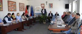 Szerokie ujęcie sali konferencyjnej podczas oficjalnego spotkania. W pomieszczeniu ustawione są drewniane stoły w kształcie podkowy, przy których siedzi kilkanaście osób – kobiety i mężczyźni w średnim wieku, ubrani w stylu półformalny. Na stołach znajdują się mikrofony, butelki z wodą, dokumenty i notesy. W centrum, na końcu stołów, stoi mężczyzna w ciemnej marynarce, który czyta z kartki i wygląda na osobę prowadzącą spotkanie. Za nim widoczny jest baner z napisem „Podkarpacka Izba Rolnicza (PIR)”, a obok stoją flagi Polski i Unii Europejskiej. Na ścianie wiszą godło Polski, herb oraz krzyż. W pomieszczeniu są jasne ściany, sufitowe lampy i okna po prawej stronie, przez które wpada światło dzienne.