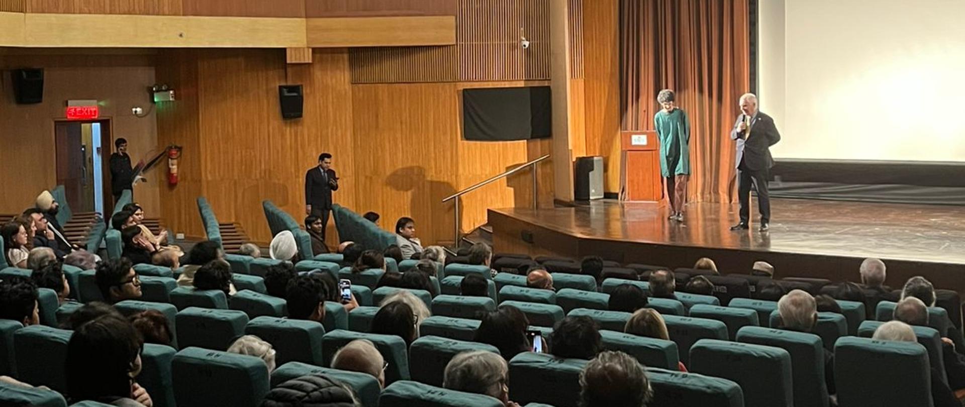 Sala kinowa pełna ludzi podczas pokazu filmu Listy z Wilczej. Na scenie przemawia Kierownik Placówki Piotr Świtalski oraz reżyser filmu Arjun Talwar