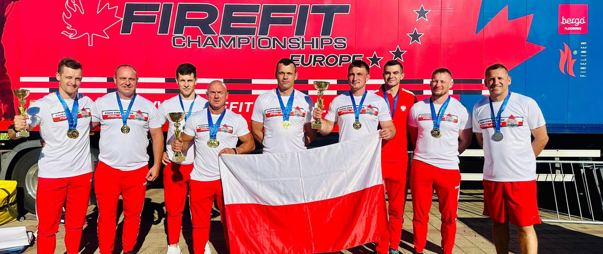 Na zdjęciu widać dużą grupę polskich strażaków pozujących na świeżym powietrzu przed czerwoną ciężarówką. Wszyscy ubrani są w białe koszulki z czerwonymi akcentami i czerwone spodnie. Dodatkowo dwaj mężczyźni trzymają polską flagę. Na ciężarówce za nimi widoczne jest logo "Firefit Championships Europe" oraz motyw liścia klonu i gwiazd.