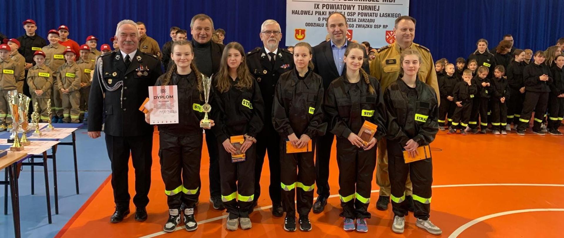 Na przedstawionym zdjęciu widać grupę osób pozujących do wspólnego zdjęcia wewnątrz hali sportowej. W centrum stoi młoda dziewczyna trzymająca dyplom i puchar, ubrana w strój przypominający mundur strażacki z widocznymi odblaskowymi paskami. Po obu stronach dziewczyny znajdują się dorośli, prawdopodobnie oficjele lub trenerzy, także ubrani w mundury. Po lewej stronie stoi mężczyzna w eleganckim mundurze, prawdopodobnie wysokiego rangą oficera straży pożarnej, uśmiechnięty i patrzący w kamerę. Po prawej stronie widać innego mężczyznę w mundurze, jak również młodą kobietę w podobnym stroju do dziewczyny z pucharem.
W tle rozciąga się długa linia dzieci i młodzieży, wszystkie w jednolitych strojach, które mogą sugerować drużynę lub uczestników jakiegoś wydarzenia. Po lewej stronie, za oficerem, stoi inny starszy mężczyzna w formalnym ciemnym mundurze. Na tle za grupą widoczny jest baner z napisem informującym o wydarzeniu sportowo-pożarniczym dla młodzieży. Puchar i dyplomy na stole wskazują, że jest to jakaś forma ceremonii wręczania nagród lub uznania osiągnięć. Atmosfera jest uroczysta, a uczestnicy wydają się być dumni i zadowoleni.