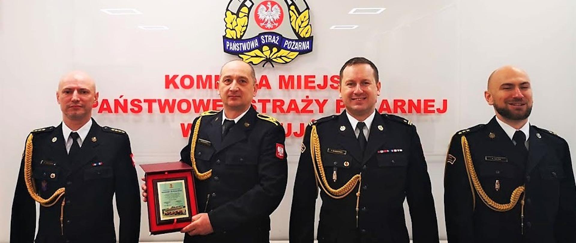 Czterech strażaków w mundurach wyjściowych en face, w sali konferencyjnej KM PSP w Świnoujściu.
Od lewej st. kpt. Marek Wieczorek, st. ogn. Marek Bernach – prezentujący pamiątkowy ryngraf, mł. bryg. Tomasz Samborski, kpt. Konrad Gajos.
