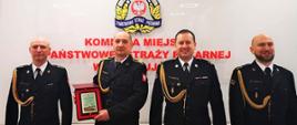 Czterech strażaków w mundurach wyjściowych en face, w sali konferencyjnej KM PSP w Świnoujściu.
Od lewej st. kpt. Marek Wieczorek, st. ogn. Marek Bernach – prezentujący pamiątkowy ryngraf, mł. bryg. Tomasz Samborski, kpt. Konrad Gajos. 