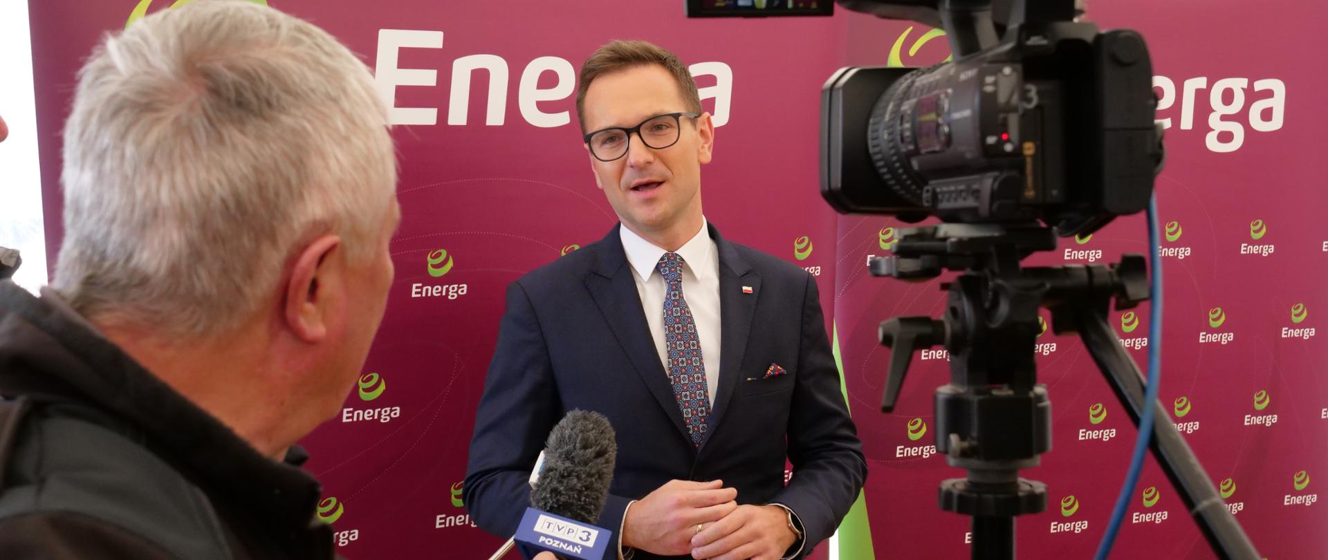 Na zdjęciu stoi wiceminister Waldemar Buda na tle bordowej planszy, obok stoi dziennikarz z mikrofonem i kamera.