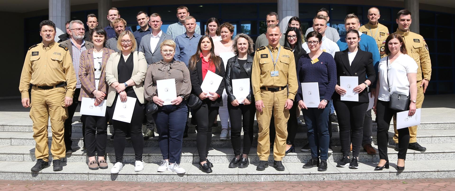 Grupowe zdjęcie uczestników szkolenia (25 osób) w towarzystwie Komendanta oraz Zastępcy Komendanta Centralnej Szkoły PSP oraz dwóch funkcjonariuszy CS PSP
