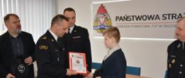 Zastępca komendanta powiatowego PSP w Radomsku wręcza dyplom Komendanta Głównego PSP chłopcu. W tle komendant powiatowy PSP w Radomsku 