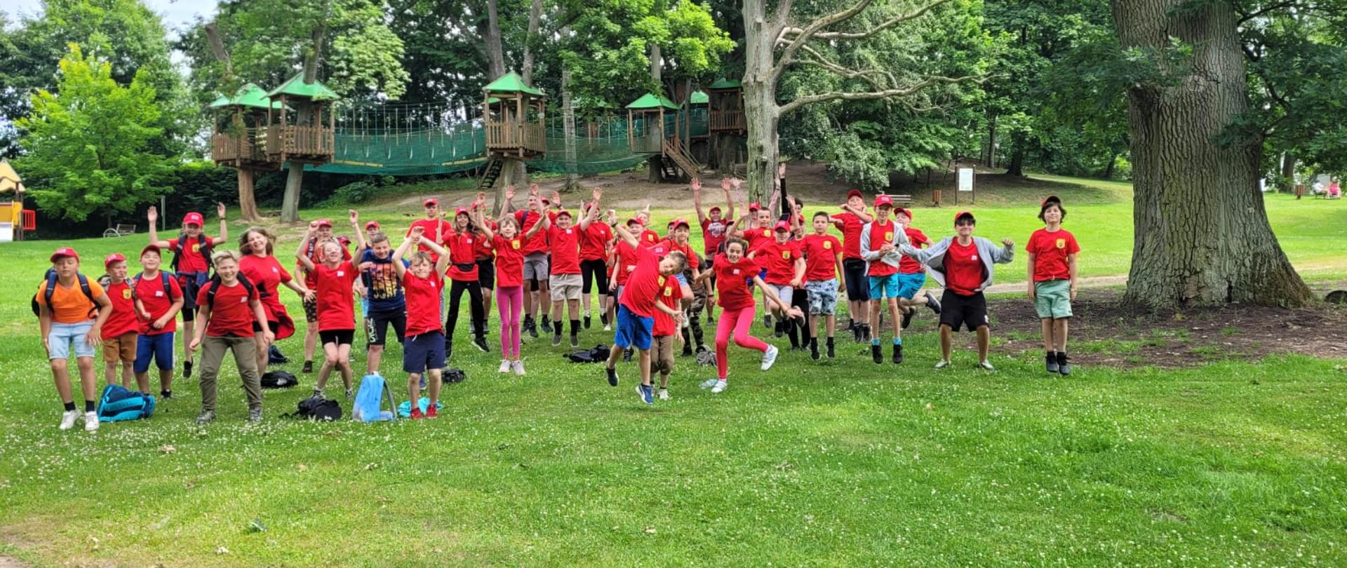 Około 50 osób - dzieci i opiekunowie w czerwonych koszulkach i czapkach podskakują z uniesionymi rękoma. W tle plac zabaw. Całość na zielonej trawie pomiędzy drzewami