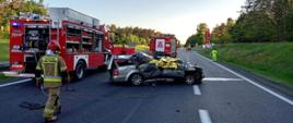 Stojący na jezdni autostrady rozbity samochód osobowy. Obok niego i za nim samochody specjalne straży pożarnej. Widoczny umundurowany strażak PSP. W tle stojący na jezdni śmigłowiec LPR.