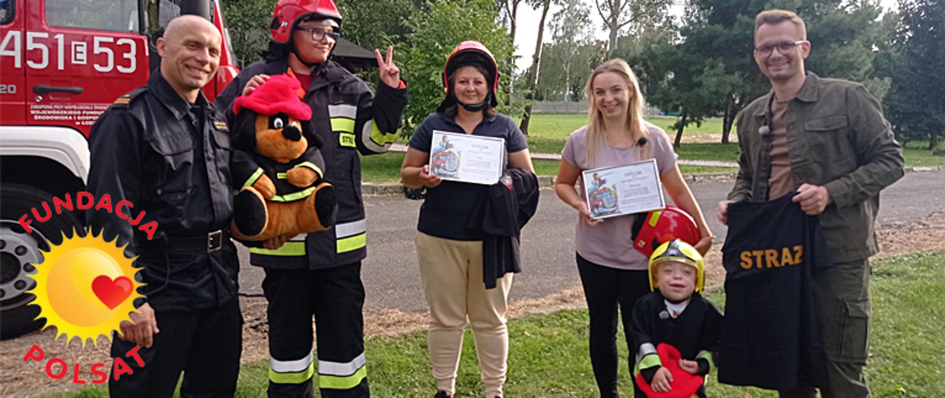 Sześć osób pozuje do fotografii, dwie kobiety trzymają przed sobą pamiątkowe dyplomy uczestników zdjęcia, którzy ubrani są w ubrania bojowe strażaków, dwóch chłopców i jedna kobieta maja na głowie hełm strażacki, druga kobieta trzyma pod pachą hełm strażacki, mężczyzna obok niej trzyma koszulkę barwy czarnej z napisem STRAŻ skierowanym do przodu, po lewej stronie fotografii stoi umundurowany strażak w ubranie barwy czarnej i trzyma pluszową maskotkę ubrana w strażackie ubranko z hełmem w kolorze czerwonym, w tle pojazd ciężarowy straży pożarnej z numerem bocznym 451E53 barwy czerwonej oraz drzewa, wszyscy stoją na zielonej trawie, słońce za chmurami, bezwietrznie. Po lewej stronie fotografii w dolnym narożniku logo fundacji Polsatu w postaci żółtego słoneczka z czerwonym serduszkiem na jego prawym boku oraz napisem barwy czerwonej ułożonym w górnej i dolnej części słoneczka wygiętych na półokrągło o treści FUNDACJA POLSAT