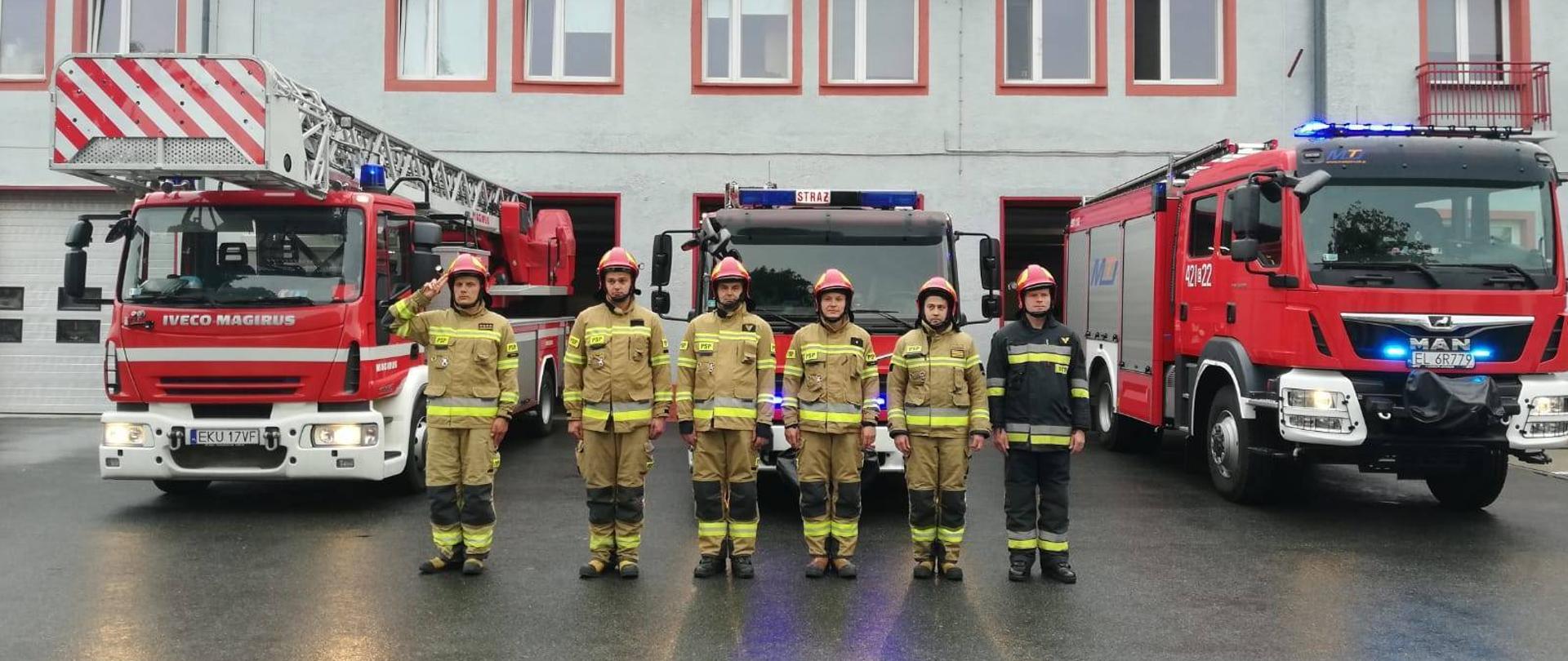 Strażacy stoją na baczność przed budynkiem Komendy PSP. Strażacy ubrani w żółte i czarne ubrania specjalne. Za nimi 3 wozy strażackie na włączonych sygnałach świetlnych i dźwiękowych.