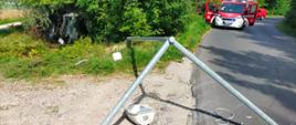 Na zdjęciu widać miejsce wypadku. W zaroślach po lewej stronie wystaje przód uszkodzonego pojazdu znajdującego się w pozycji na boku. Uszkodzona lampa częściowo przewróciła się na jezdnię. W tle widać czerwone samochody strażackie