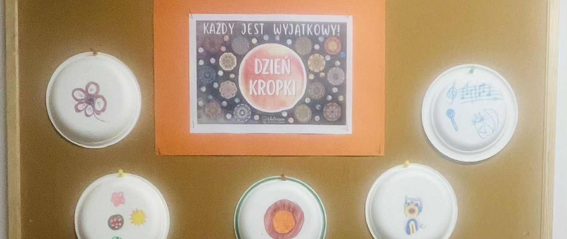 Na tablicy korkowej umieszczone zostały białe, okrągłe, papierowe talerzyki na których umieszczono rysunki np. kwiatki, globus, kotek. W górnej części tablicy, po środku umieszczono kartkę z napisem: Każdy jest wyjątkowy. Dzień Kropki.