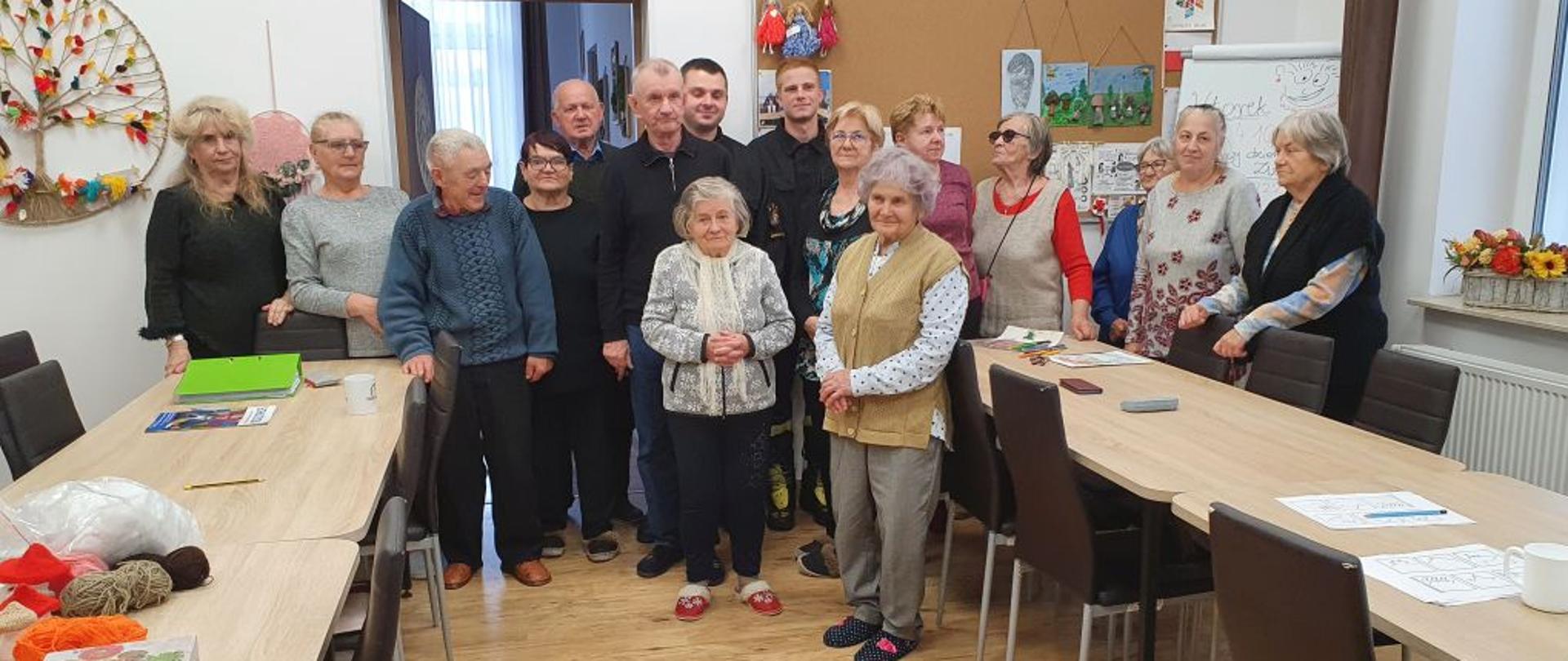 Na zdjęciu widać dwóch strażaków w ubraniach koszarowych którzy stoją w obecności czternastu seniorów. Wszyscy znajdują się w sali gdzie widoczne są stoły, krzesła, tablica korkowa różne ozdoby, a nad drzwiami wisi godło Polski.
