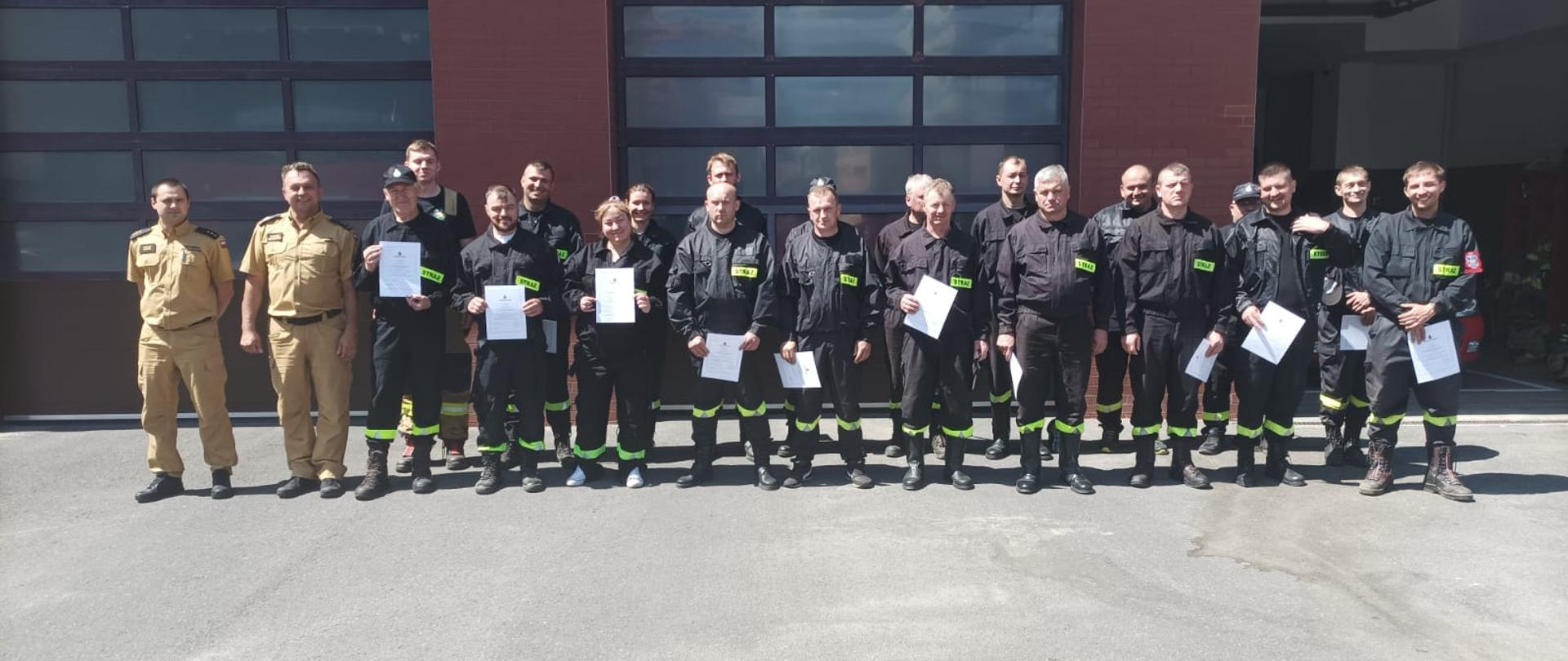 grupowe zdjęcie 20 strażaków ubranych w czarne ubrania strażackie stoja w dwóch rzędach przed budynkiem komendy obok nich stoji dwóch strażaków w ubraniach koloru piaskowego 