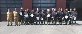 grupowe zdjęcie 20 strażaków ubranych w czarne ubrania strażackie stoja w dwóch rzędach przed budynkiem komendy obok nich stoji dwóch strażaków w ubraniach koloru piaskowego 
