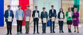 Na środku sali gimnastycznej stoi w rzędzie dziewięć młodych osób, pokazują trzymane w rękach dyplomy, za nimi na ścianie polska flaga.