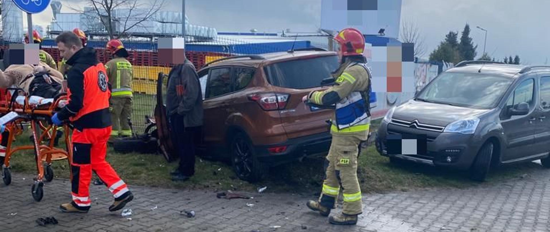 rozbity samochód koloru brązowego stoi na poboczu drogi z boku osoba postronna i jedna osoba na noszach pod opieka ratownika medycznego wokół samochodu strażacy. Za samochodem widoczny zakład produkcyjny, obok samochodu brązowego inny samochód