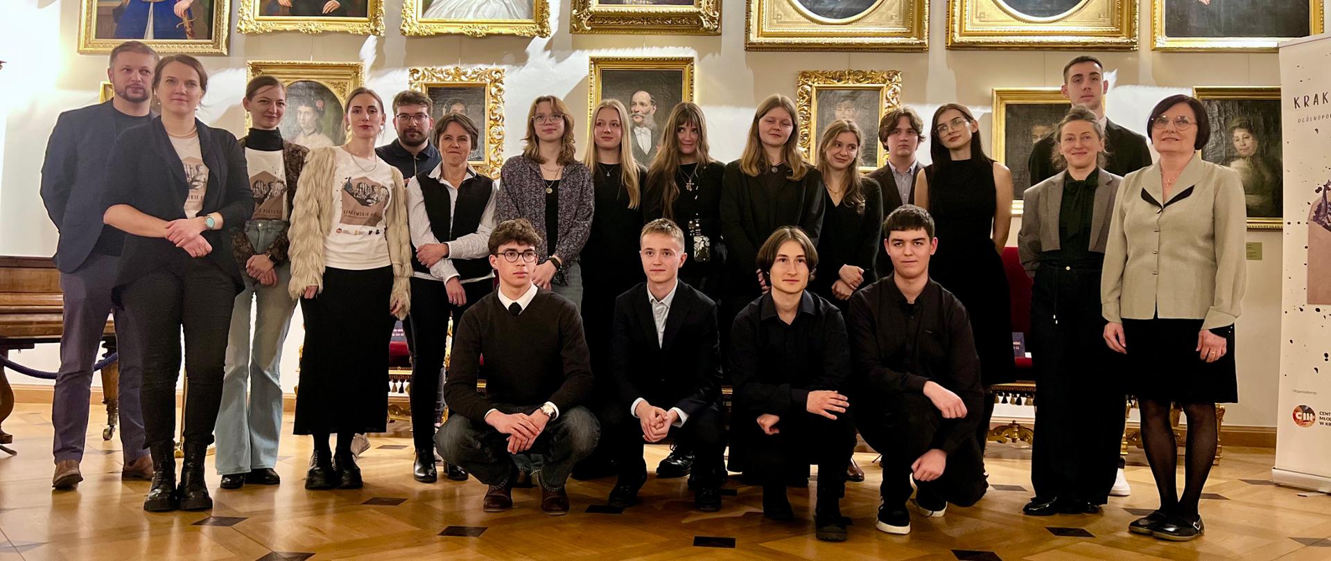 Grupa około trzydziestu osób stoi i klęczy we wnętrzu eleganckiej galerii sztuki. Za nimi wiszą liczne, bogato zdobione portrety w złotych ramach. Większość osób pozuje do wspólnego zdjęcia, część ubrana jest formalnie, a część bardziej swobodnie. 