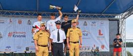 Trzech zawodników na podium, przed nimi stoi 3 strażaków PSP w mundurach