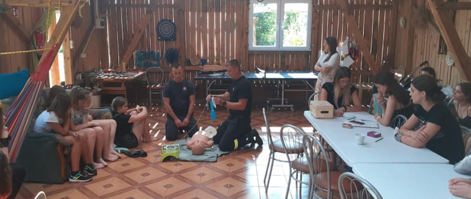 Dwóch strażaków pokazuje uczestnikom obozu harcerskiego zasady udzielania pierwszej pomocy przy użyciu defibrylatora. 