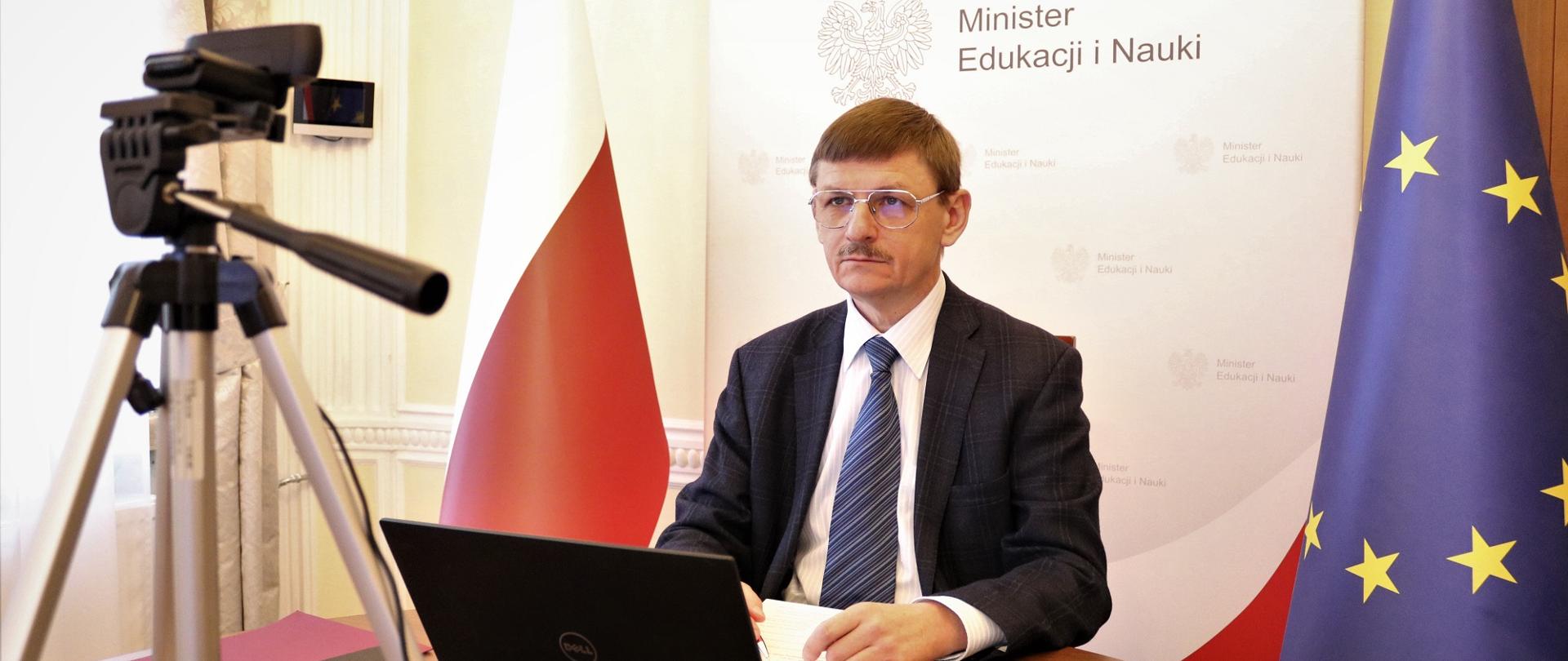 Prof. Grzegorz Wrochna siedzi przy biurku, przed nim laptop i kamera na statywie.