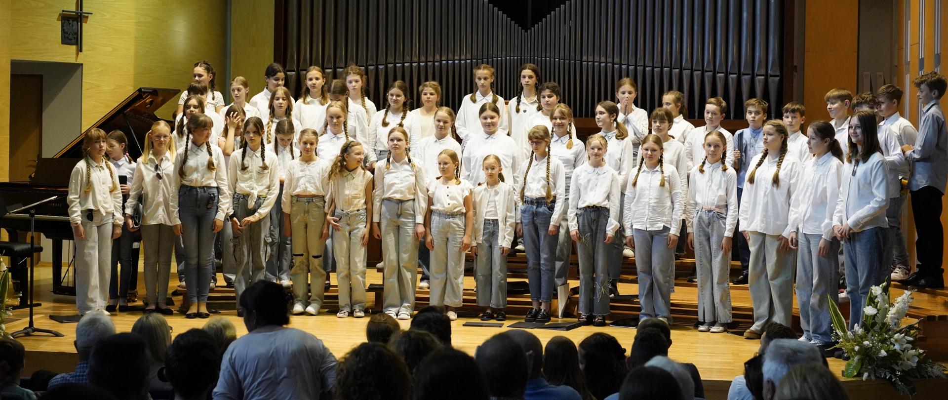 Chór dziecięcy na scenie sali koncertowej stoi w trzech rzędach. Na pierwszym planie publiczność. Za chórem organy