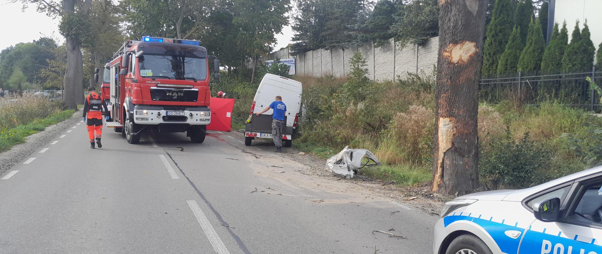 Samochód dostawczy na poboczu za uszkodzonym przydrożnym drzewem. Z lewej strony samochód strażacki.