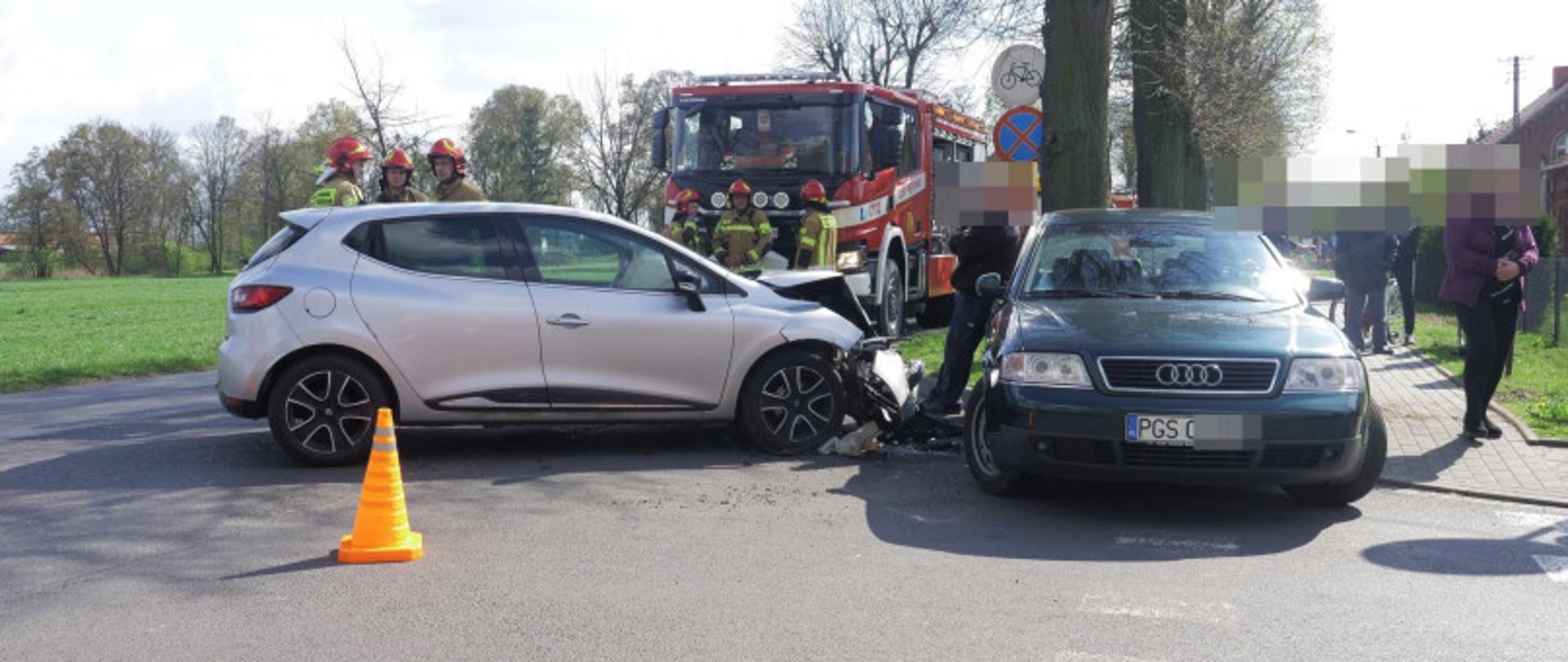 Na skrzyżowaniu dróg doszło do zderzenia dwóch pojazdów osobowych. W tle stoją strażacy oraz samochód pożarniczy.