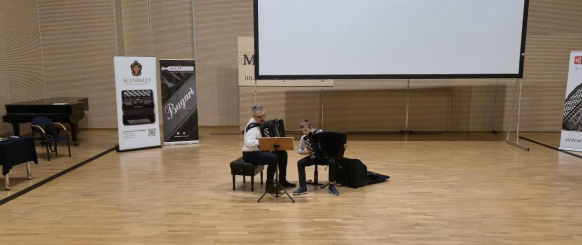 Zdjęcie przedstawia dwóch muzyków grających na akordeonach na scenie w auli, w tle widoczny jest duży ekran projekcyjny oraz roll-upy reklamowe producentów instrumentów.
