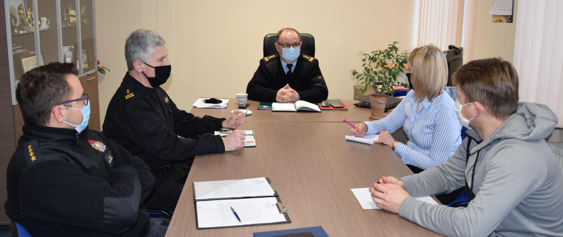  Pięć osób zasiadających przy stole konferencyjnym w pokoju komendanta. Trzech strażaków, mężczyzna i kobieta. 