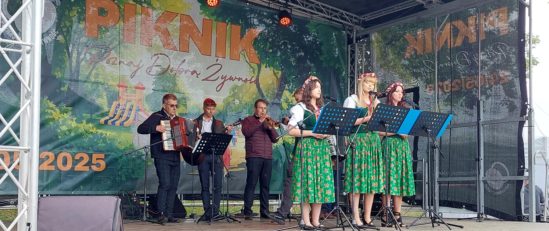 Scena na której stoją przy mikrofonach trzy młode kobiety w ludowych strojach i kwietnych wiankach na głowie. Za nimi stoją czterej mężczyźni grający na instrumentach ludowych.