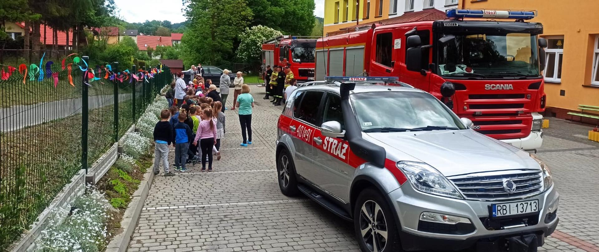Na zdjęciu widać samochód dowódcy oraz dwa wozy bojowe Państwowej Straży Pożarnej stojące przed przedszkolem oraz dzieci ustawione przed nimi w parach.