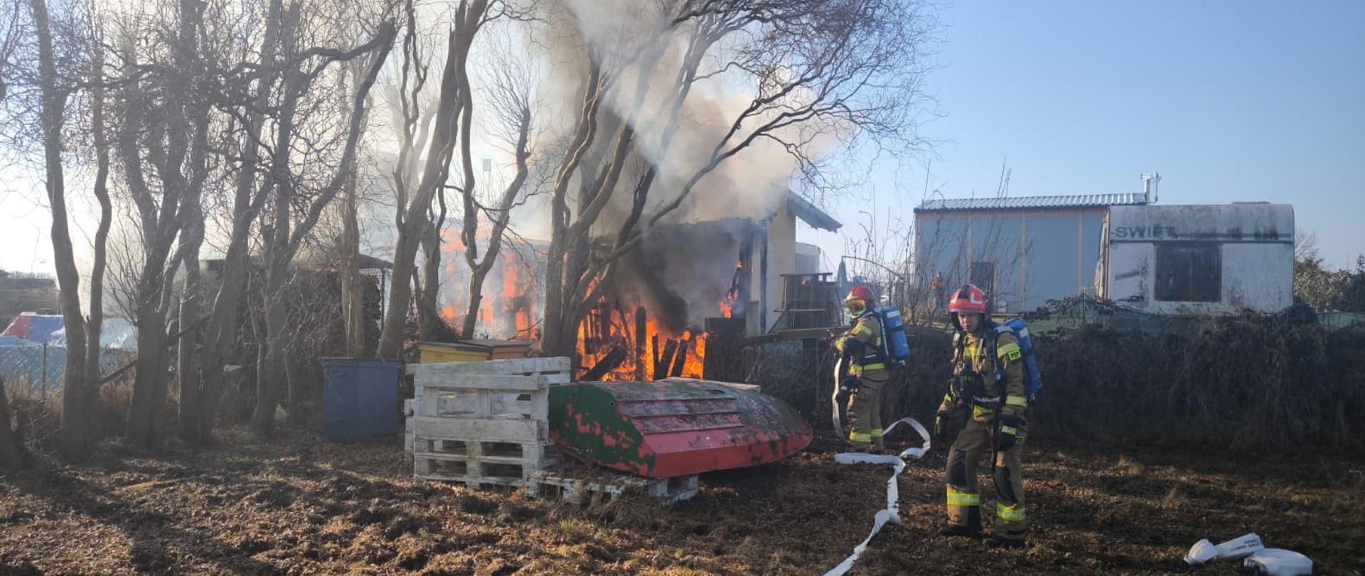 Zdjęcie przedstawia pożar budynku letniskowego. Na zdjęciu widoczni strażacy w trakcie akcji gaśniczej ubrani w piaskowe mundury z czerwonymi hełmami.