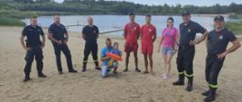 Wspólne patrolowanie akwenów. Na plaży w półkolu stoi trzech strażaków, dwóch policjantów,dwóch ratowników WOPR i trzy osoby cywilne, w tym mężczyzna z dzieckiem na rękach. W tle akwen z pomostami, drzewa i krzewy. Strażacy występują w umundurowaniu koszarowym.